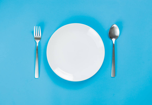 Ein weißer Teller mit Löffel und Gabel auf blauem Hintergrund - Foto, Bild