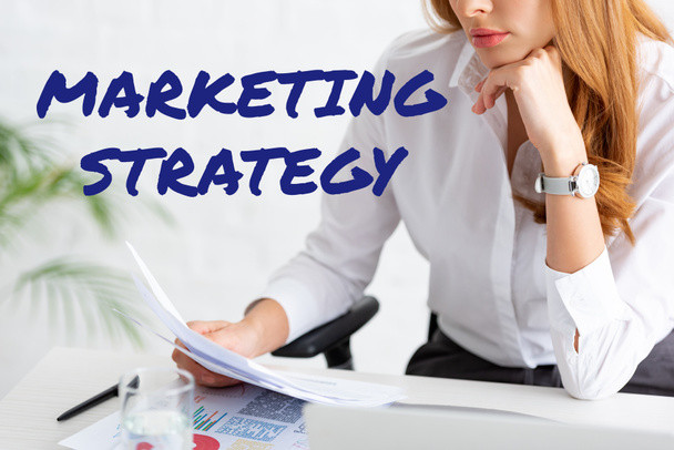 Vista cortada de empresária segurando dossiê perto de gráficos e laptop na mesa, ilustração estratégia de marketing
 - Foto, Imagem