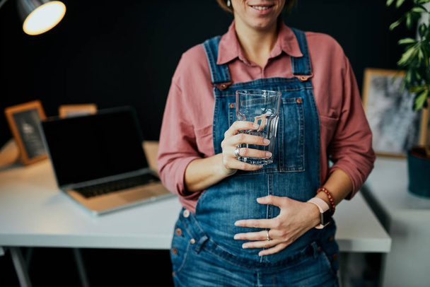 Kobieta w ciąży stojąca w swoim gabinecie, dotykająca brzucha i pijąca słodką wodę. - Zdjęcie, obraz