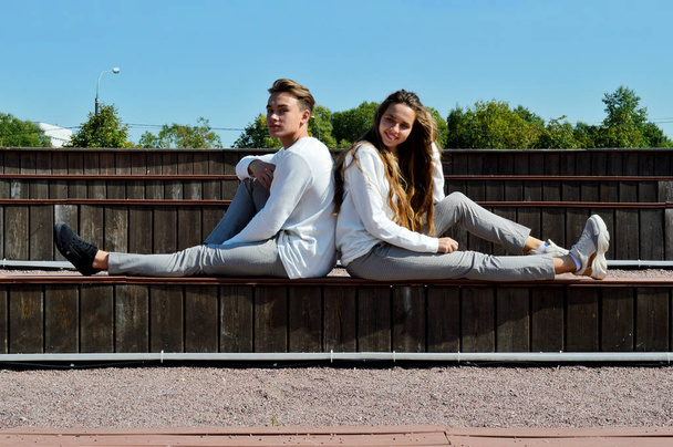 Een jong meisje en een jonge man in dezelfde kleren zittend op een houten bank. - Foto, afbeelding