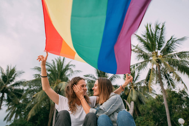 Dos mujeres con bandera de arco iris en la playa sobre un fondo de palma
 - Foto, imagen