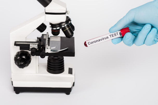 abgeschnittene Ansicht eines Wissenschaftlers, der eine Probe mit Coronavirus-Test-Schriftzug in der Nähe des Mikroskops hält, isoliert auf weiß  - Foto, Bild