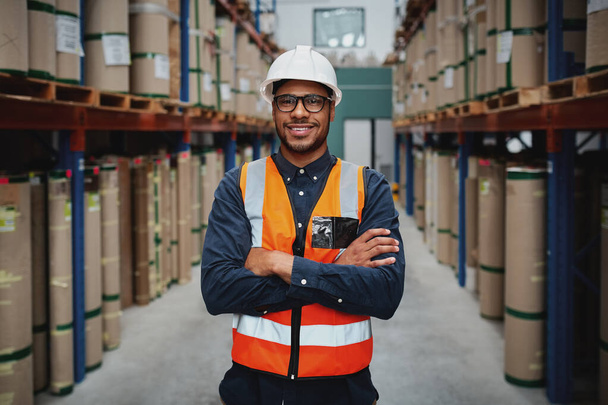 Portrait smiling worker wearing orange vest and white helmet with arms crossed in a large warehouse smiling - Foto, Bild