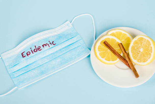 bovenaanzicht van schotel met kaneelstokjes en citroenen bij medisch masker met epidemische letters op blauw - Foto, afbeelding