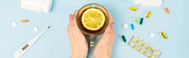 panoramic shot of woman holding cup of tea with sliced lemon near pills and digital thermometer on blue  - Photo, Image