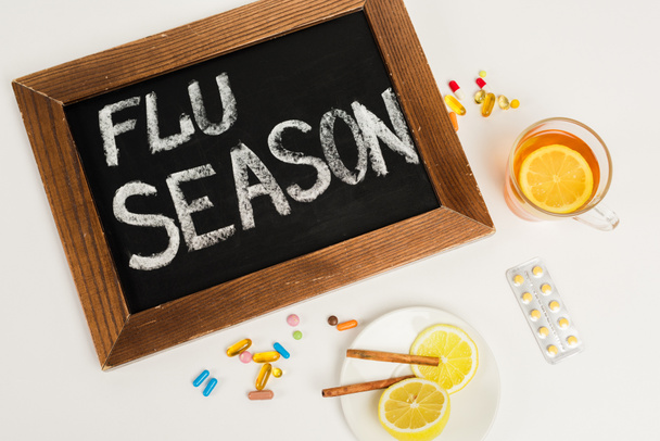 Kreidetafel mit Grippesaison-Schriftzug in der Nähe von Pillen und Tee mit Zitrone auf Weiß  - Foto, Bild