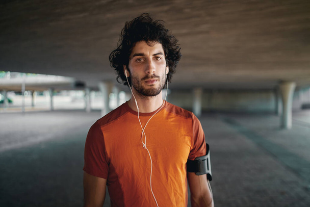 Portrait of serious fit young man with earphones in his ear standing under the bridge looking at the camera - Foto, imagen