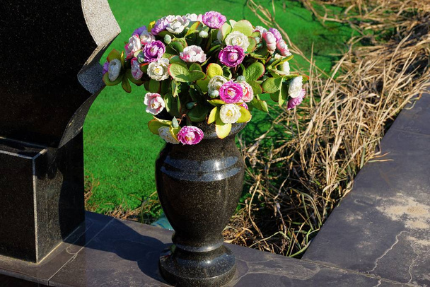 een zwarte marmeren vaas met een boeket van gekleurde kunstbloemen staat op een stenen plaat op een begraafplaats - Foto, afbeelding