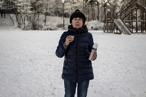 De winter. Een dame met een hoed, een blauwe sportjas en jeans houdt een thermoskan en een mok tegen de achtergrond van een besneeuwd bos - Foto, afbeelding