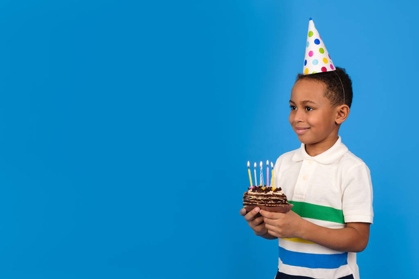 Happy African American boy with funny little party cone on head celebrates birthday holding chocolate cake in hands with burning candles and rejoicing in holiday on blue background. Birthday concept - Фото, зображення