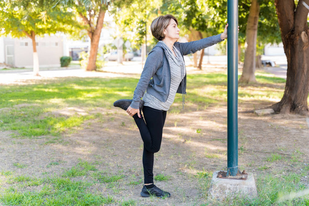 Longitud completa de la mujer mayor estirando su pierna mientras se apoya en el poste en el parque
 - Foto, imagen