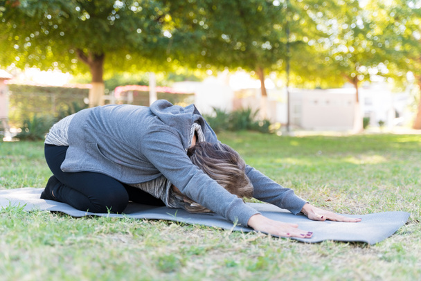 Flexibele oudere vrouw die haar rug uitrekt tijdens het trainen op de mat in het park - Foto, afbeelding
