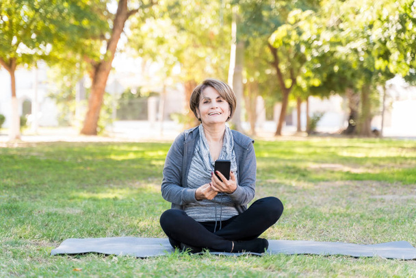 Portrait of happy retired elderly woman holding mobile phone while sitting with cross legged on yoga mat - Photo, Image
