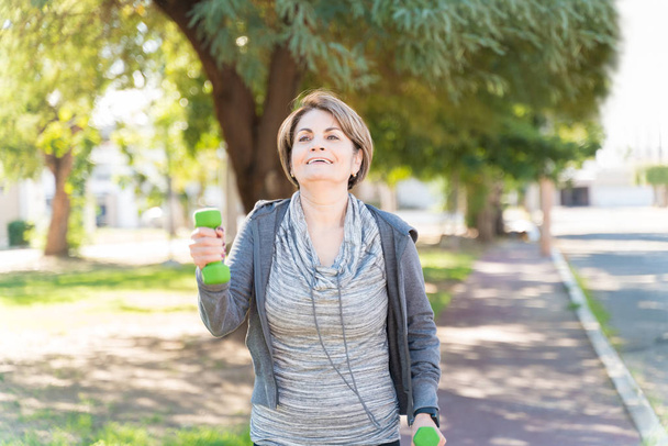 Smiling active elderly Caucasian woman walking while lifting dumbbells in city - Photo, Image