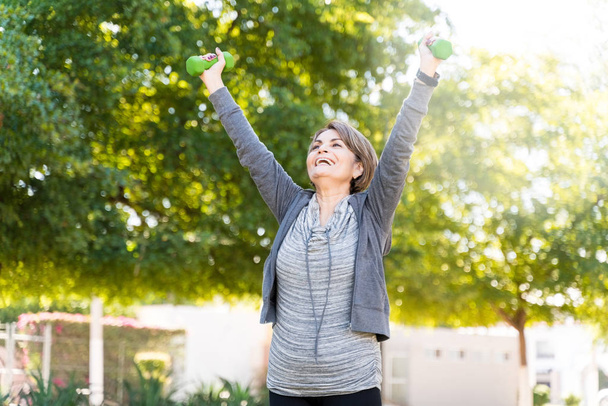 Mujer mayor alegre haciendo ejercicio de fitness mientras levanta pesas en el parque durante el entrenamiento
 - Foto, Imagen