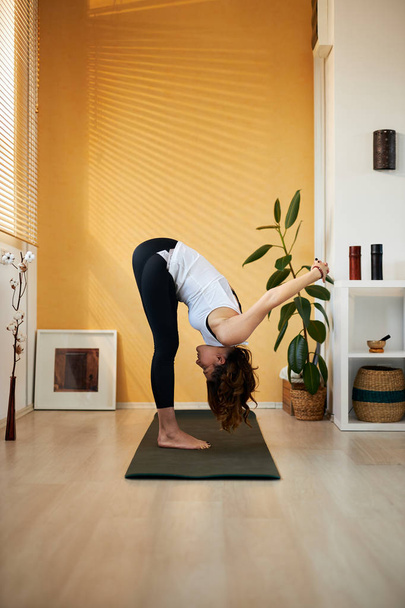 Вид збоку приваблива брюнетка середнього віку, що стоїть на килимку для йоги в Стендінг Форвард Бенд з плечем відкривачка йога позує вдома вранці
. - Фото, зображення