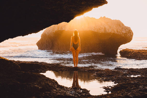 Viaggiatore donna in costume da bagno in posa al tramonto vicino oceano e roccia
 - Foto, immagini