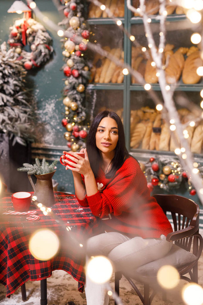 young beautiful brunette girl in a red sweater and white pants drinks tea on a street in Paris against the background of a window of a bread shop - Foto, imagen