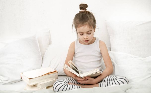 Cute little girl reading a book on the bed in the bedroom. - Foto, afbeelding