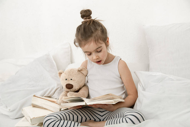 Χαριτωμένο κοριτσάκι που διαβάζει ένα βιβλίο στο κρεβάτι στην κρεβατοκάμαρα.. - Φωτογραφία, εικόνα