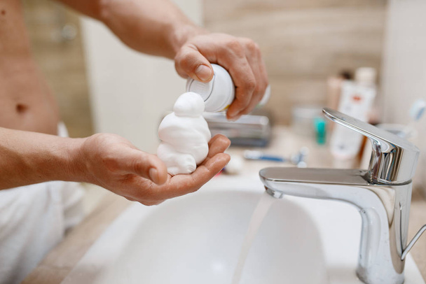 Man squeezes out shaving foam on hand in bathroom, routine morning hygiene. Male person at the sink performs skin and body treatment procedures - Fotó, kép