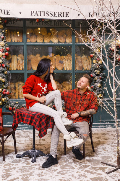 Young beautiful couple, a brunette girl in a red sweater and white pants, a man in a red shirt are sitting on the streets of Paris against the background of a window of a bakery shop - Photo, Image