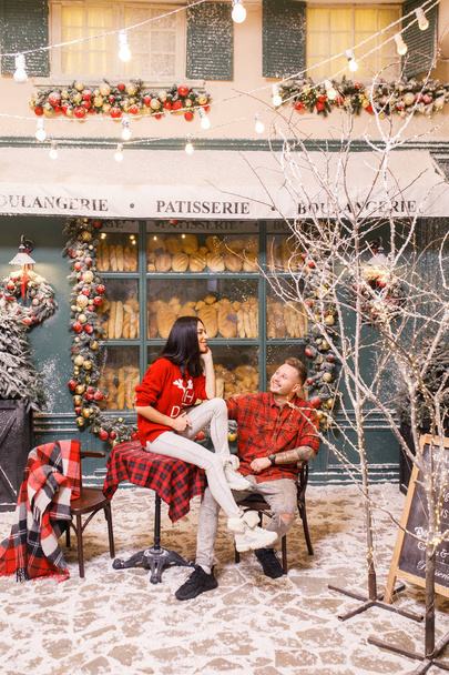 Young beautiful couple, a brunette girl in a red sweater and white pants, a man in a red shirt are sitting on the streets of Paris against the background of a window of a bakery shop - Фото, зображення