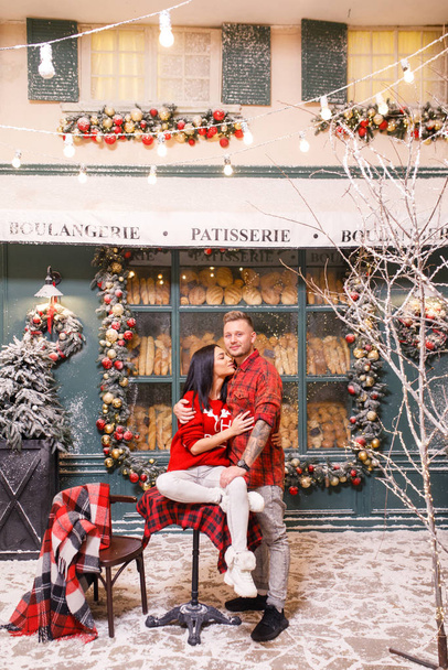 Young beautiful couple, a brunette girl in a red sweater and white pants, a man in a red shirt are sitting on the streets of Paris against the background of a window of a bakery shop - 写真・画像