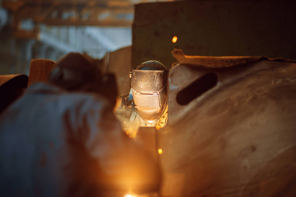 Két maszkos hegesztő készül arra, hogy fémszerkezettel dolgozzon a gyárban, hegesztési készséggel. Fémfeldolgozó ipar, acéltermékek ipari gyártása - Fotó, kép