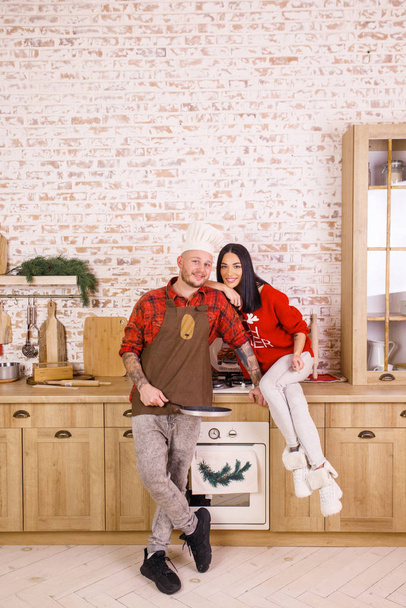 Joven hermosa pareja, morena en un suéter rojo y pantalones blancos, un hombre con una camisa roja en la cocina preparando la comida juntos
 - Foto, imagen