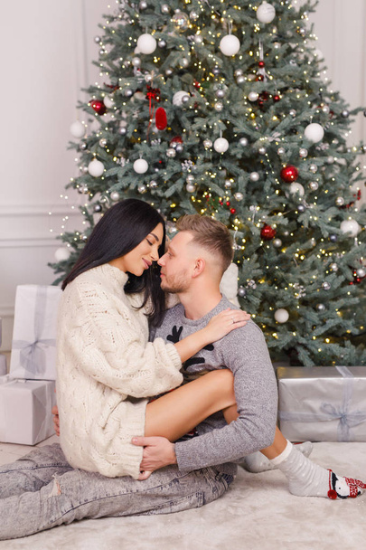 young beautiful couple, brunette girl in a white sweater, a blond man in a gray sweater sitting near a Christmas tree - Fotografie, Obrázek