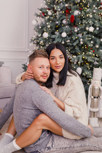 young beautiful couple, brunette girl in a white sweater, a blond man in a gray sweater sitting near a Christmas tree - Фото, изображение
