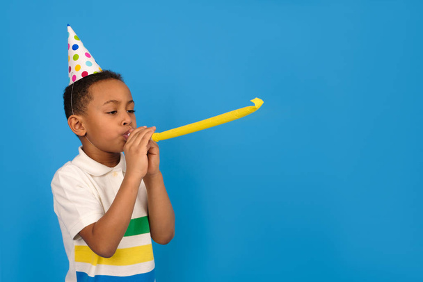 Happy African-American boy celebrates his birthday blowing a whistle on a blue background. Holiday and party concept with space for text. - Foto, Imagen