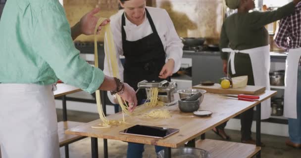 Side view of a senior Caucasian man and a Caucasian female chef during a cookery class in a restaurant kitchen, the chef explaining and helping the man to roll dough through a pasta machine, in slow motion - Video