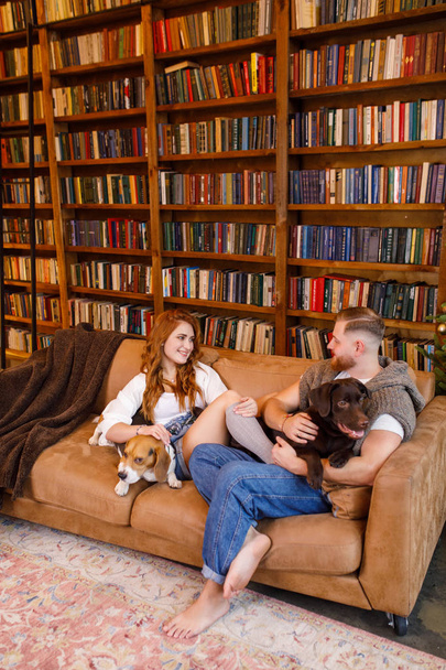 young beautiful couple sitting on the bed with two dogs on the background of the library and books in the apartment in the loft style - Foto, Imagen