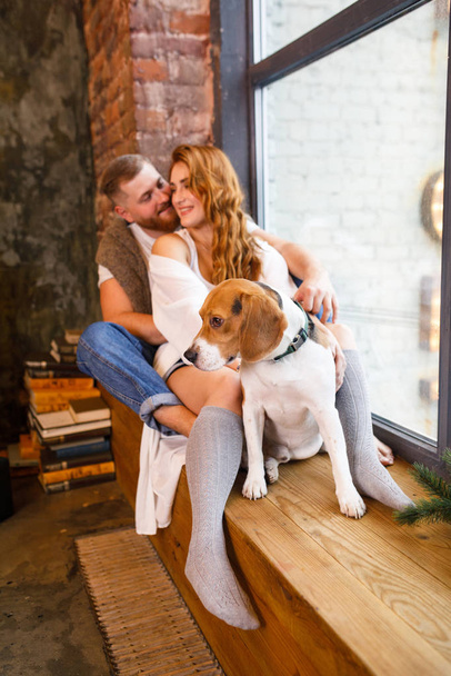 young beautiful couple is sitting at the window with two dogs in a loft apartment - 写真・画像