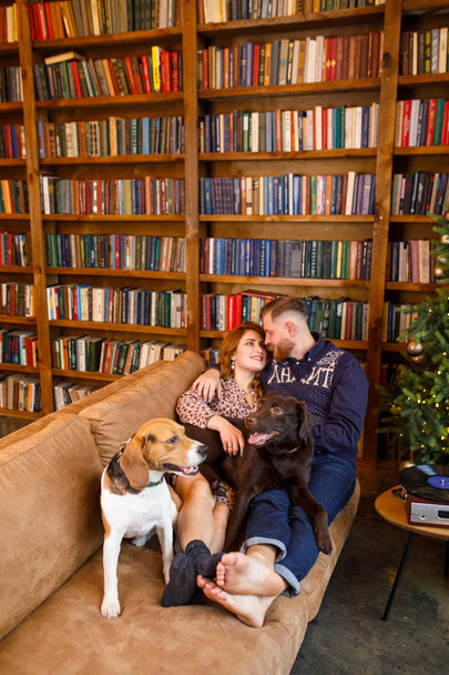 young beautiful couple sitting on the bed with two dogs on the background of the library and books in the apartment in the loft style - Fotoğraf, Görsel