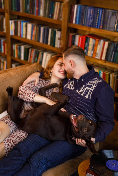 young beautiful couple sitting on the bed with two dogs on the background of the library and books in the apartment in the loft style - Valokuva, kuva