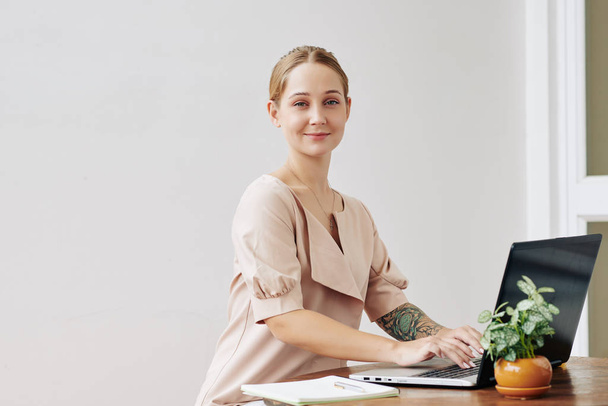 Горизонтальный портрет современной деловой женщины с татуировкой на руке, смотрящей в камеру во время работы на ноутбуке
 - Фото, изображение