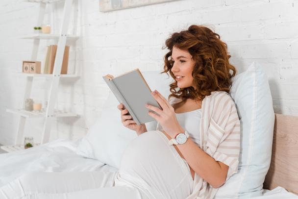 Attractive pregnant woman smiling while reading book on bed  - 写真・画像