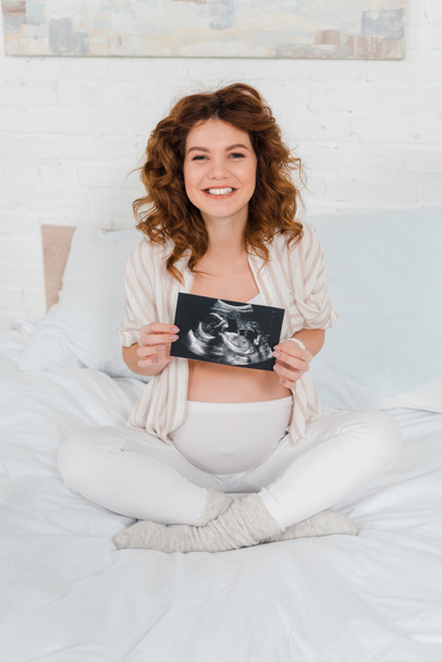 Pregnant woman smiling at camera while holding ultrasound scan of baby on bed  - Foto, Bild
