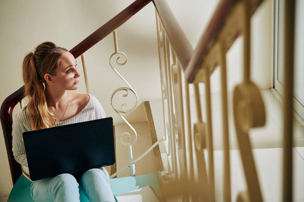 Горизонтальный снимок стильной молодой женщины, сидящей с ноутбуком на лестнице и смотрящей в сторону
 - Фото, изображение