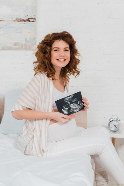 Beautiful pregnant woman smiling at camera while holding ultrasound scan of baby in bedroom - Photo, Image