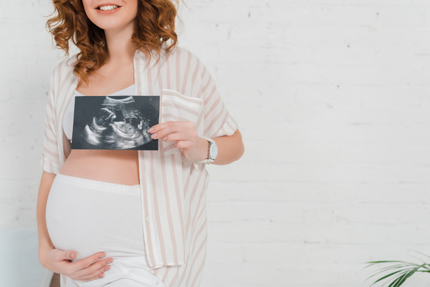 Обрезанный вид беременной девушки, показывающей ультразвуковое сканирование ребенка и трогательный живот
 - Фото, изображение