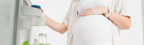Ausgeschnittene Ansicht einer schwangeren Frau, die Kühlschranktür auf weißem Hintergrund öffnet, Panoramaaufnahme - Foto, Bild