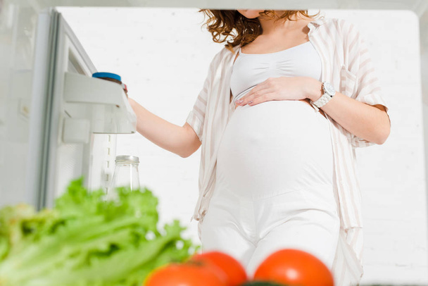 Обрезанный вид беременной женщины с рукой на животе открытия холодильника со свежими овощами на белом фоне
 - Фото, изображение