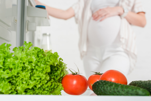 Selective focus of pregnant woman opening fridge with fresh vegetables isolated on white - Фото, изображение
