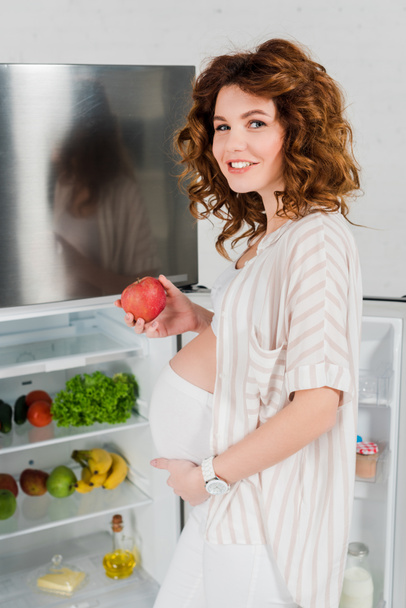 Side view of pregnant woman smiling at camera while holding apple near open fridge with fresh vegetables and fruits - Fotó, kép