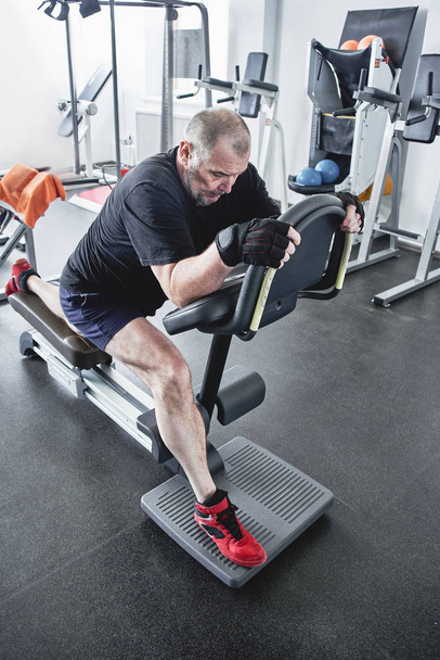 Adult gray-haired man trains on fitness equipment in the gym, pumps legs and arms muscles, loses weight. Concept of healthy lifestyle in old age - Photo, image