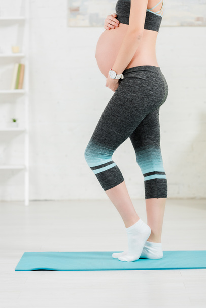 Cropped view of pregnant woman touching belly while standing on fitness mat in living room - Foto, Imagem